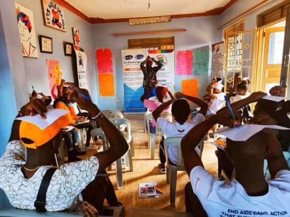 Classroom of students facing their teacher and drawing on a sheet of paper on top of their heads. Credit: Let's Walk Uganda